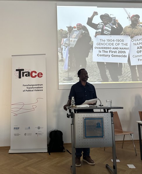 Ein Mann steht in der Mitte des Bildes vor einem Pult und spricht, während sein Blick nach vorne gerichtet ist. Links hinter ihm steht ein Banner mit TraCe-Logo, daneben ist ein Bild von demonstrierenden Menschen an die Wand projiziert.