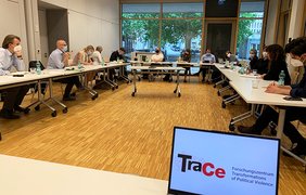 Teilnehmer:innen des Kolloquiums sitzen um einen großen Tisch in U-Form. Vorne im Bild ist das TraCe-Logo auf einem Laptopbildschirm zu sehen.