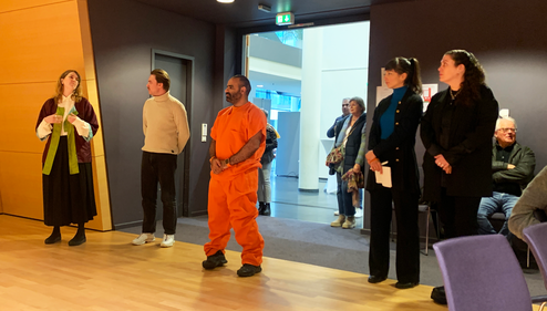 Five people are standing in front of a door. One man is wearing an orange prison uniform.