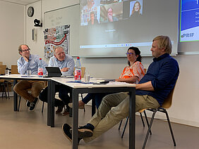Four people are sitting at a table, talking to one another. In the background, there is a Zoom panel showing participants joining the call remotely.