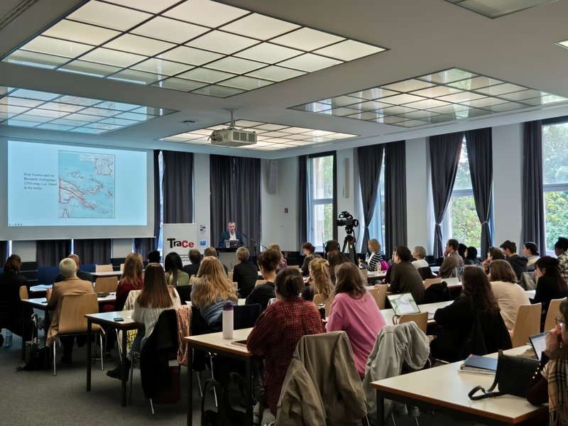 Ein Raum voller Menschen, die nach vorne auf eine Leinwand sehen, auf der eine Landkarte abgebildet ist. Daneben steht ein Mann an einem Podium und hält einen Vortrag. 