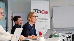 A woman sits at a conference table talkin. Next to her, two people sit around the table listening. A TraCe Roll-Up can be seen in the background
